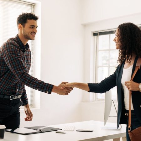 Businessman greeting a woman entrepreneur at his table by shaking hand. Businesswoman carrying office bag shaking hand with a business colleague at his office.