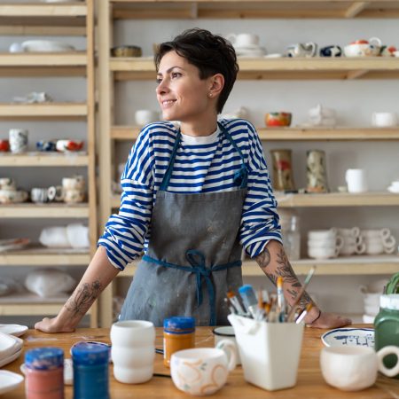 Calm young female owner in pottery workshop looking at window and smiling, plans projects. Small business concept, ceramics studio. Successful italian woman entrepreneur with tattoos at workplace.